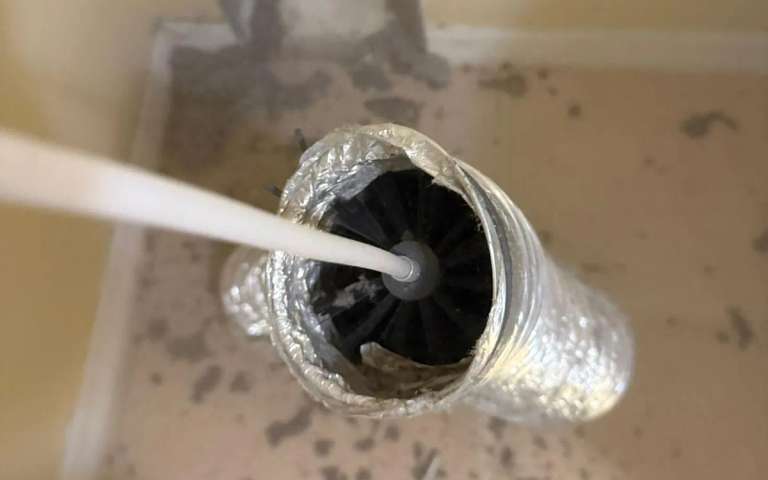 Un cepillo rotativo en un cable blanco dentro de un ducto de secadora de aluminio cubierto de pelusa, con pelusa esparcida por el piso
