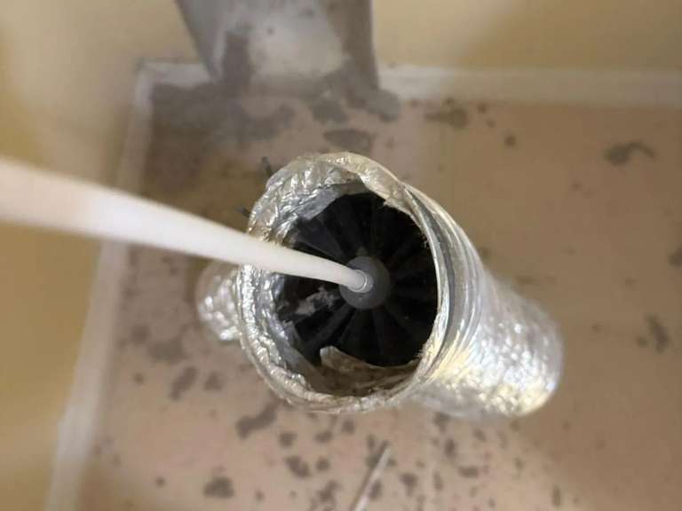 Un cepillo rotativo en un cable blanco dentro de un ducto de secadora de aluminio cubierto de pelusa, con pelusa esparcida por el piso