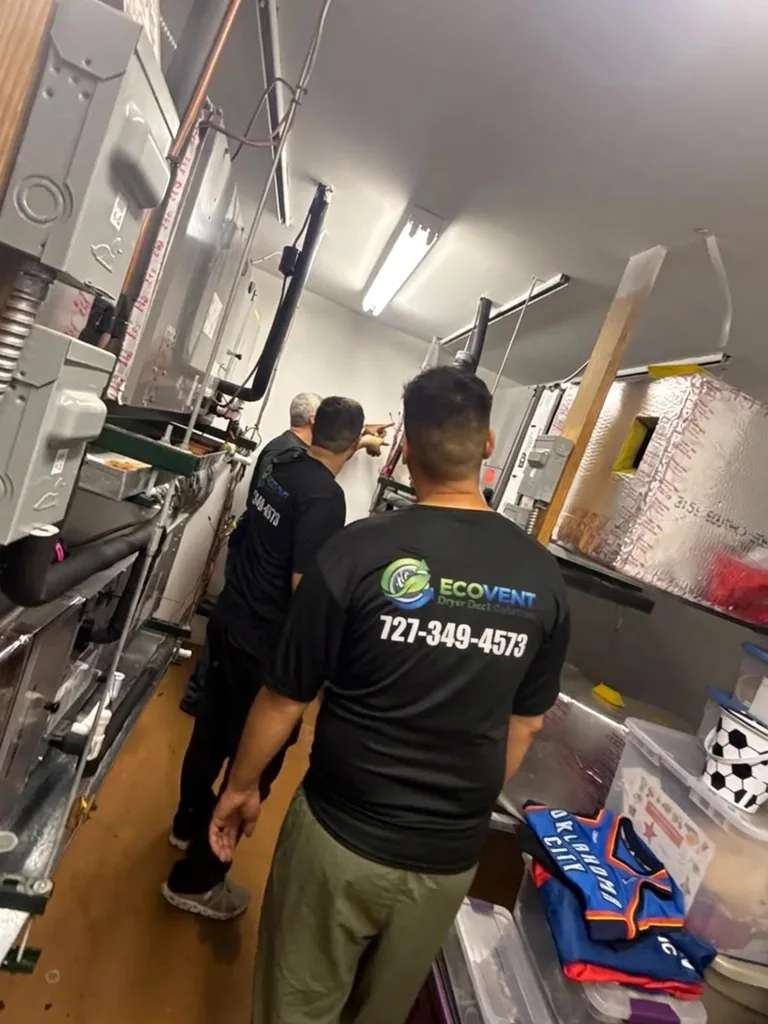 Tres miembros del equipo de Ecovent en un cuarto de máquinas con unidades de aire, tuberías y ductos forrados de aluminio, uno de ellos señalando el equipo