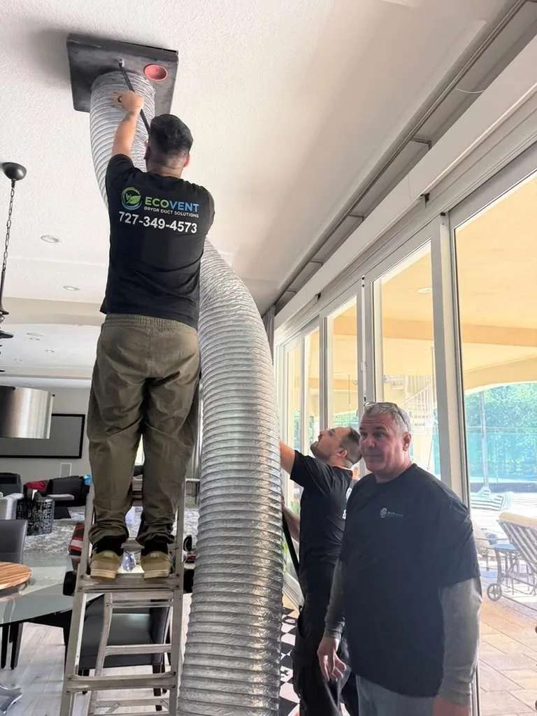Un técnico de Ecovent, subido a una escalera, sella una manguera flexible ancha de aspiración a una rejilla del techo de una sala, mientras un compañero sostiene la manguera y otro mira