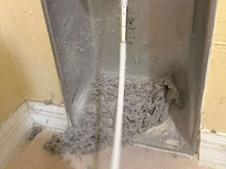 A thick pile of gray lint at the bottom of a dryer vent wall box, with a cleaning rod reaching down into it