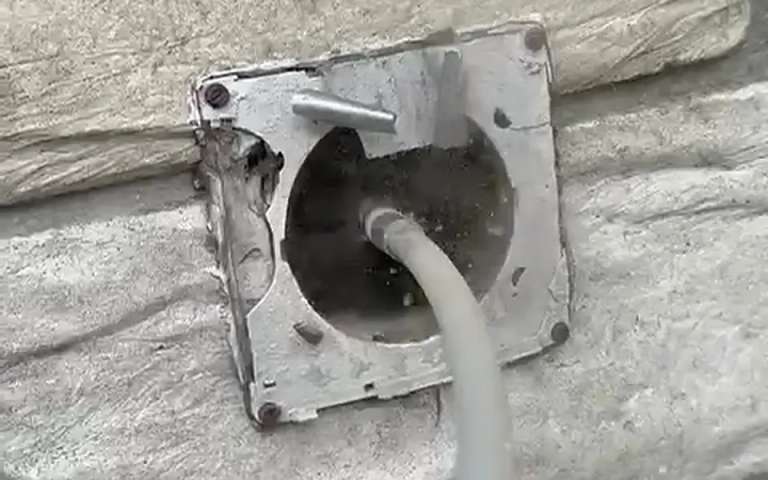 An outside dryer vent opening on a painted block wall with the flap removed and a gray cleaning cable coming out of the duct