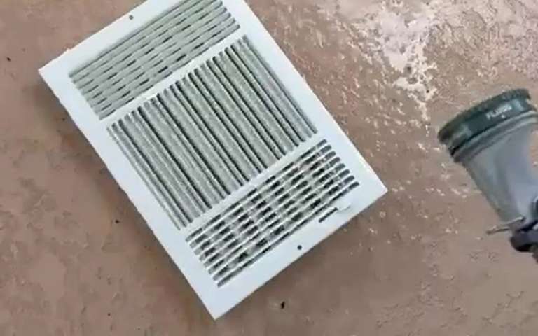 Una rejilla blanca con manchas de polvo, ya desmontada y apoyada junto a la boquilla de una manguera de aspiración