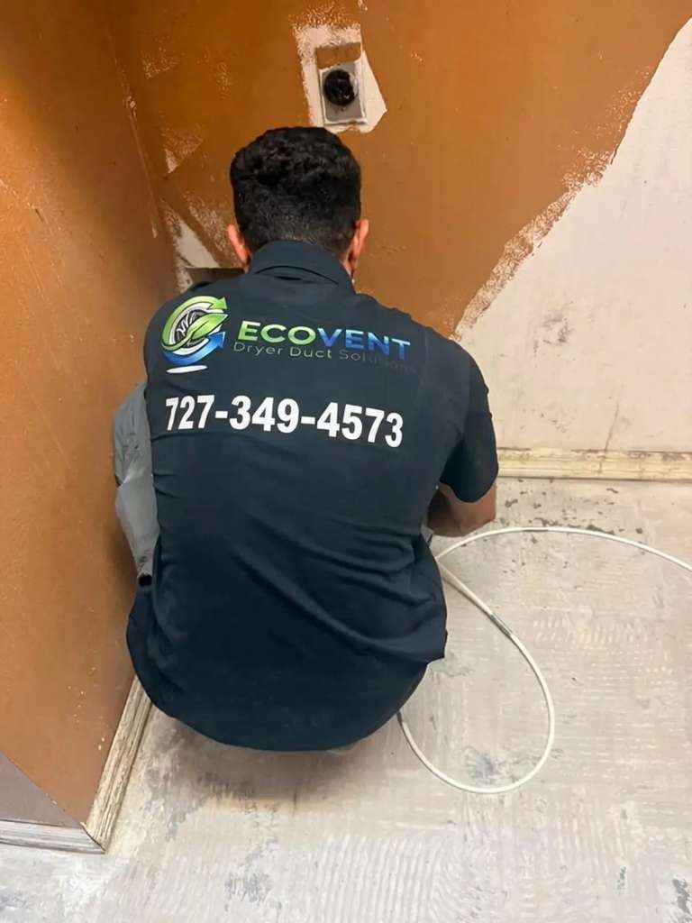Seen from behind, an Ecovent technician crouches in the corner of a laundry space and runs a white cleaning cable into the dryer vent
