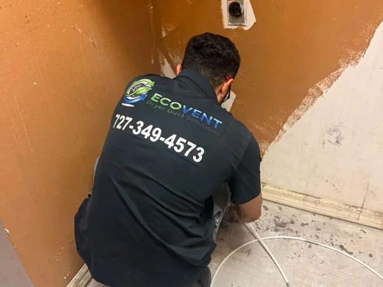 Un técnico de Ecovent, arrodillado en un hueco de lavado vacío con paredes naranjas, mete un cable blanco de limpieza en el ducto de secadora al pie de la pared