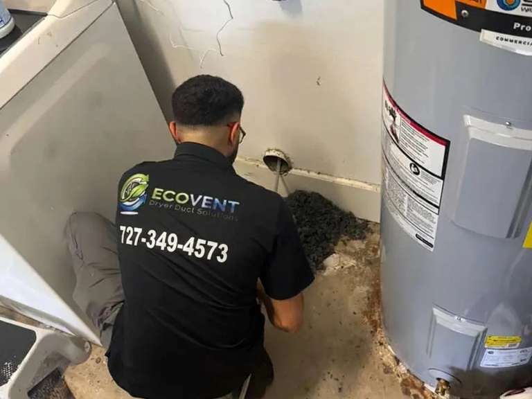 Un técnico de Ecovent, arrodillado entre la lavadora y el calentador de agua, mete un cable de limpieza en la boca del ducto de secadora de la pared, con un montón de pelusa gris ya sacada en el suelo