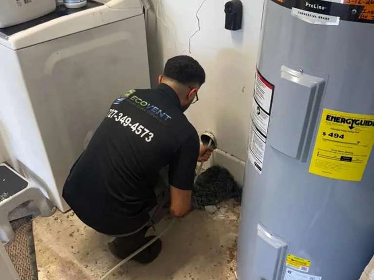 Un técnico de Ecovent, agachado junto a la lavadora y el calentador de agua, mete un cable de limpieza en la boca del ducto de secadora mientras la pelusa se amontona en el piso